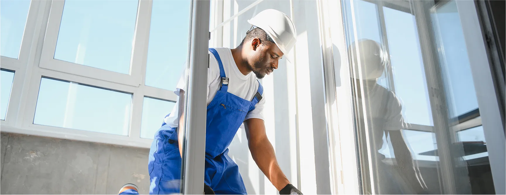 A construction worker wearing a white hard hat and blue overalls is carefully installing a large window.