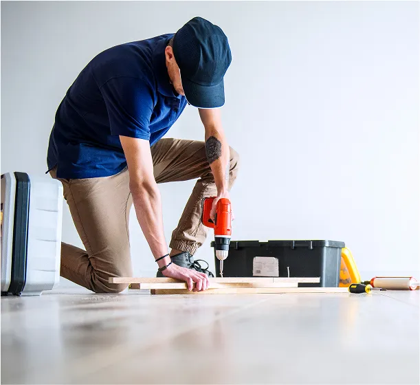 A man wearing a blue shirt and cap is kneeling on the floor while using a power drill on a wooden board.