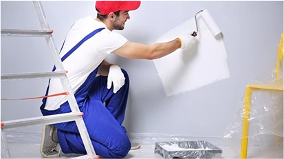 A worker in blue overalls and a red cap painting a wall with a roller.