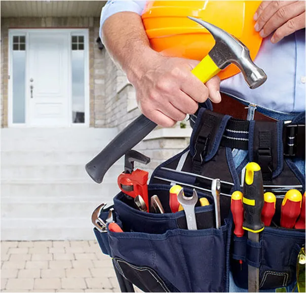 A person wearing a tool belt filled with various tools like screwdrivers, wrenches, and pliers, holding a hammer and a yellow hard hat, standing outside in front of a house.