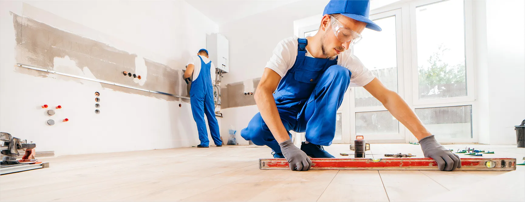 Two workers in blue overalls and caps are working indoors on home renovation. One worker is crouching and using a level tool on the floor, while the other is standing and working on the wall. The room appears to be under construction or remodeling, with exposed pipes and unfinished walls.