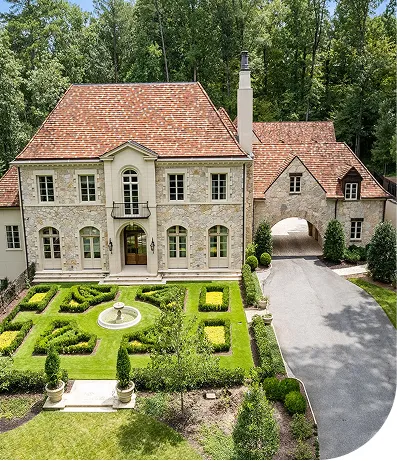 A grand two-story stone mansion with a red-tiled roof and arched windows is set in a lush, wooded area. The front yard features an elegant formal garden with manicured hedges, a central fountain, and symmetrical landscaping. A curved driveway leads to an arched entryway connecting to a side structure.