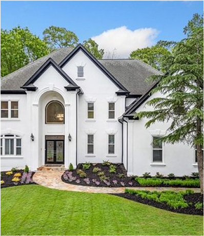 Elegant two-story white house with black trim and arched entryway, surrounded by well-manicured landscaping, vibrant flower beds, and a curved stone pathway leading to the front door. Lush green lawn and tall trees complete the scenic setting