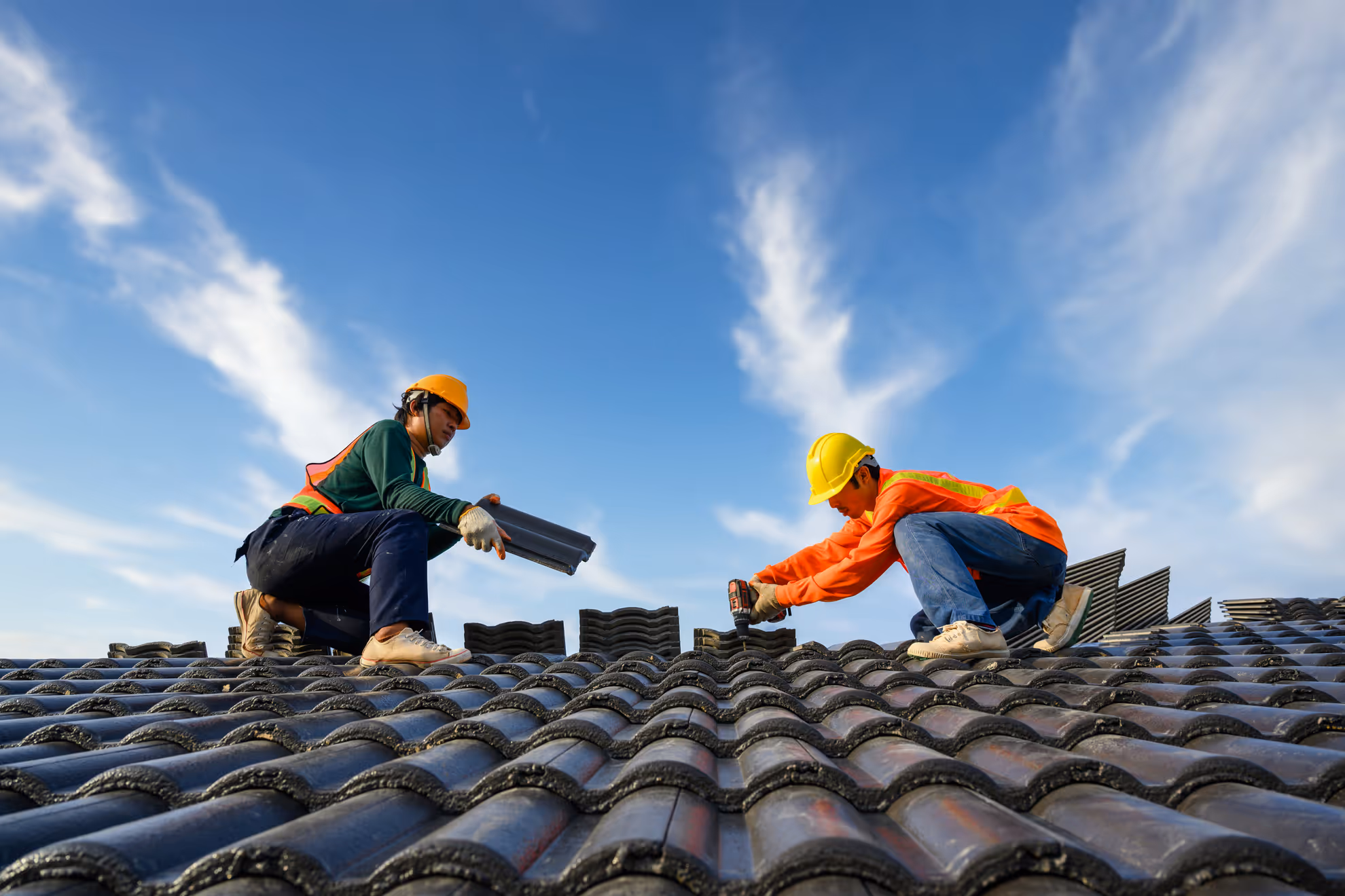 Two construction workers wearing safety gear, including helmets and reflective vests, are installing roof tiles on a building. One worker is placing a tile while the other uses a power drill to secure it. The sky is bright blue with wispy white clouds in the background.
