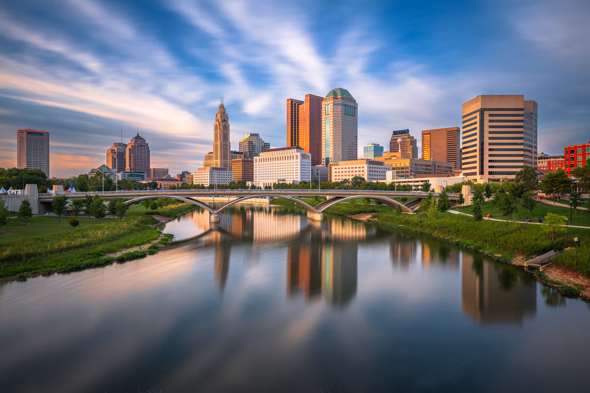 A vibrant city skyline during sunset with modern high-rise buildings reflecting on a calm river. A graceful arched bridge spans the river, connecting green parks on both sides. The sky is partly cloudy with streaks of soft, warm light illuminating the buildings and the surrounding landscape.
