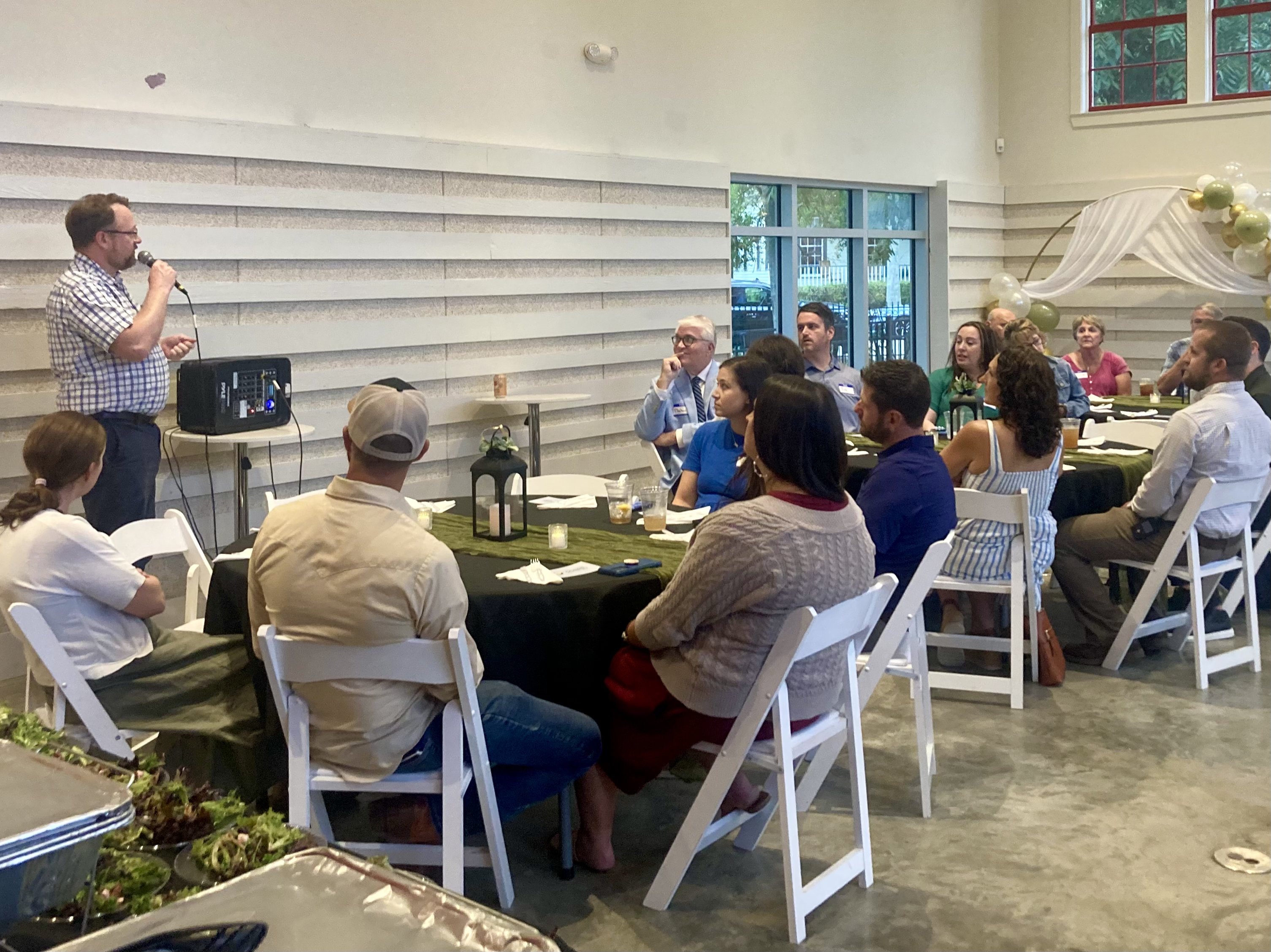 Man speaking into a microphone to a group of people seated around tables in a bright room with large windows and balloon decorations.