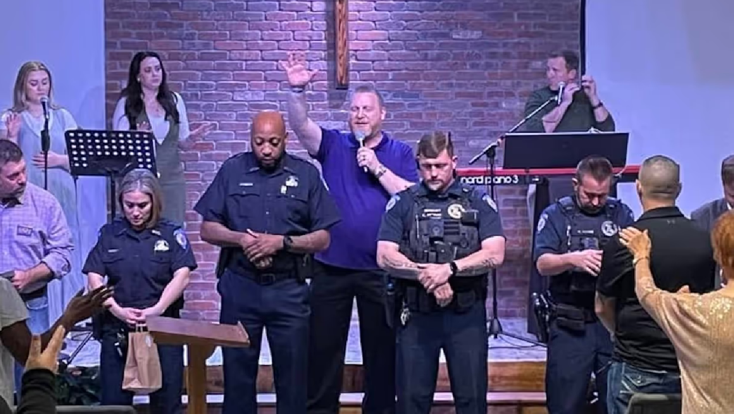 Group of police officers and civilians praying inside a church with a man raising his hand and speaking into a microphone near a brick wall with a cross.