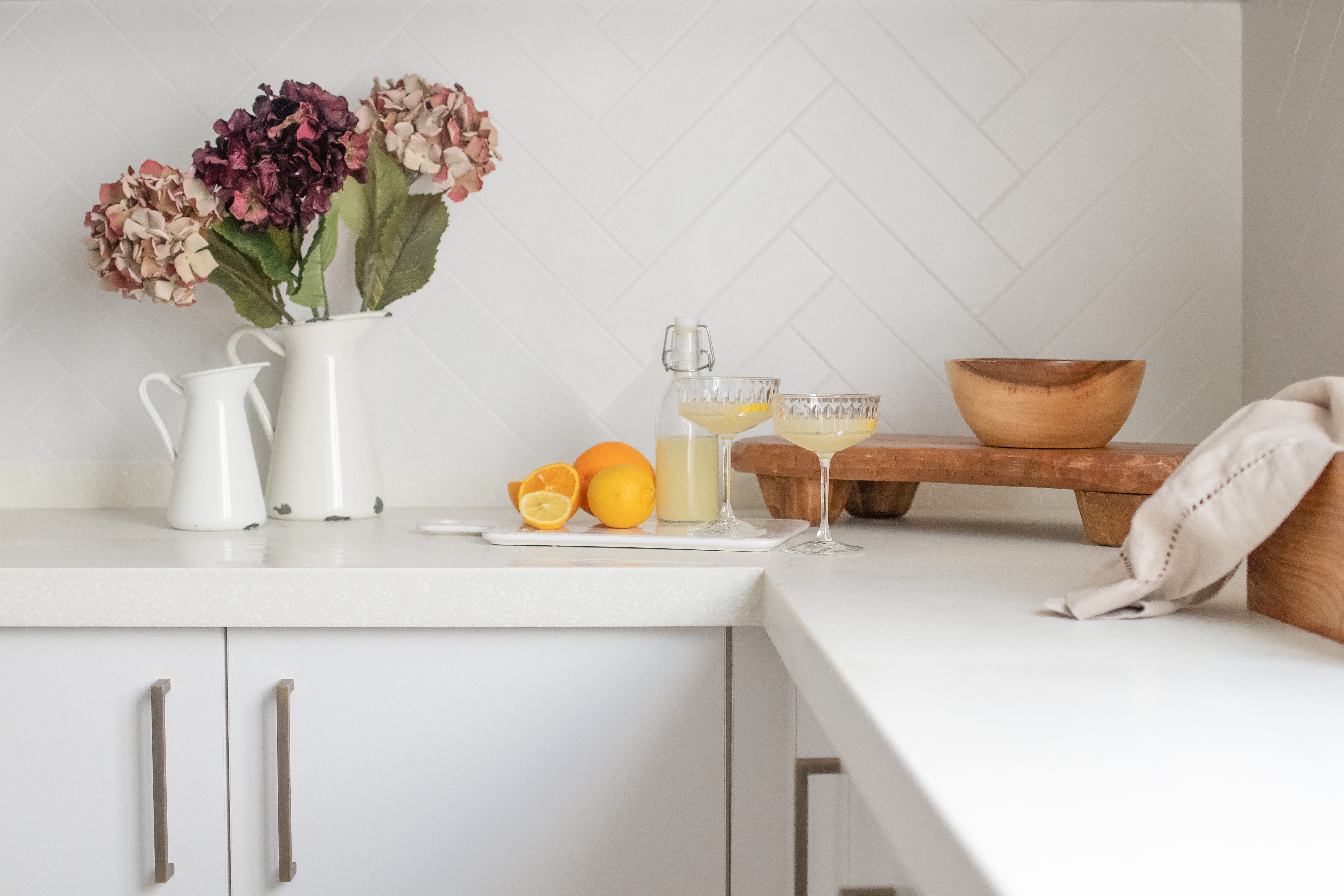Bright white kitchen worktop with citrus drinks, decorative vases, and herringbone splashback in a minimalist design.