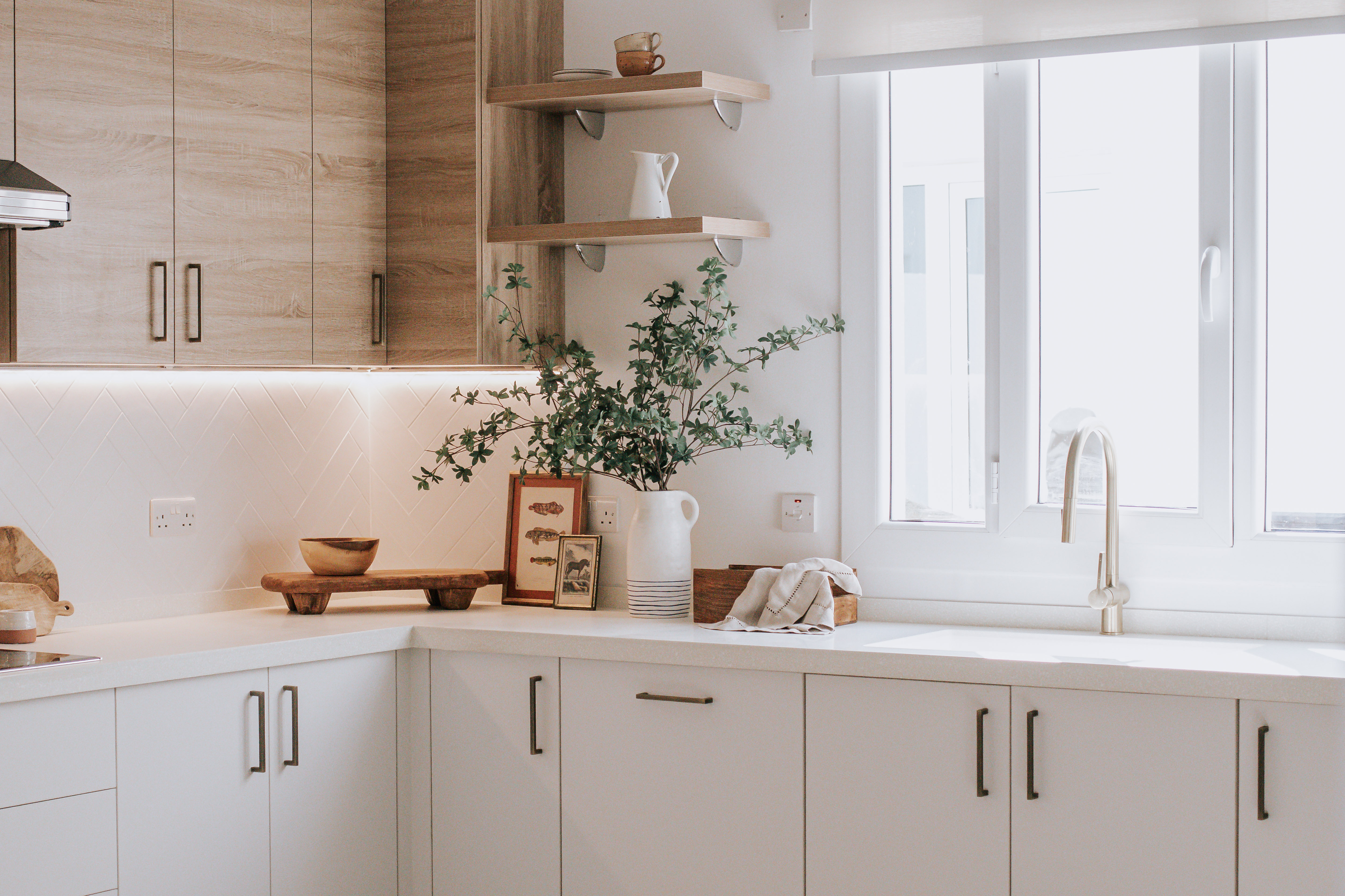 Contemporary kitchen with white units, oak wall cabinets, and styled greenery under a large window.