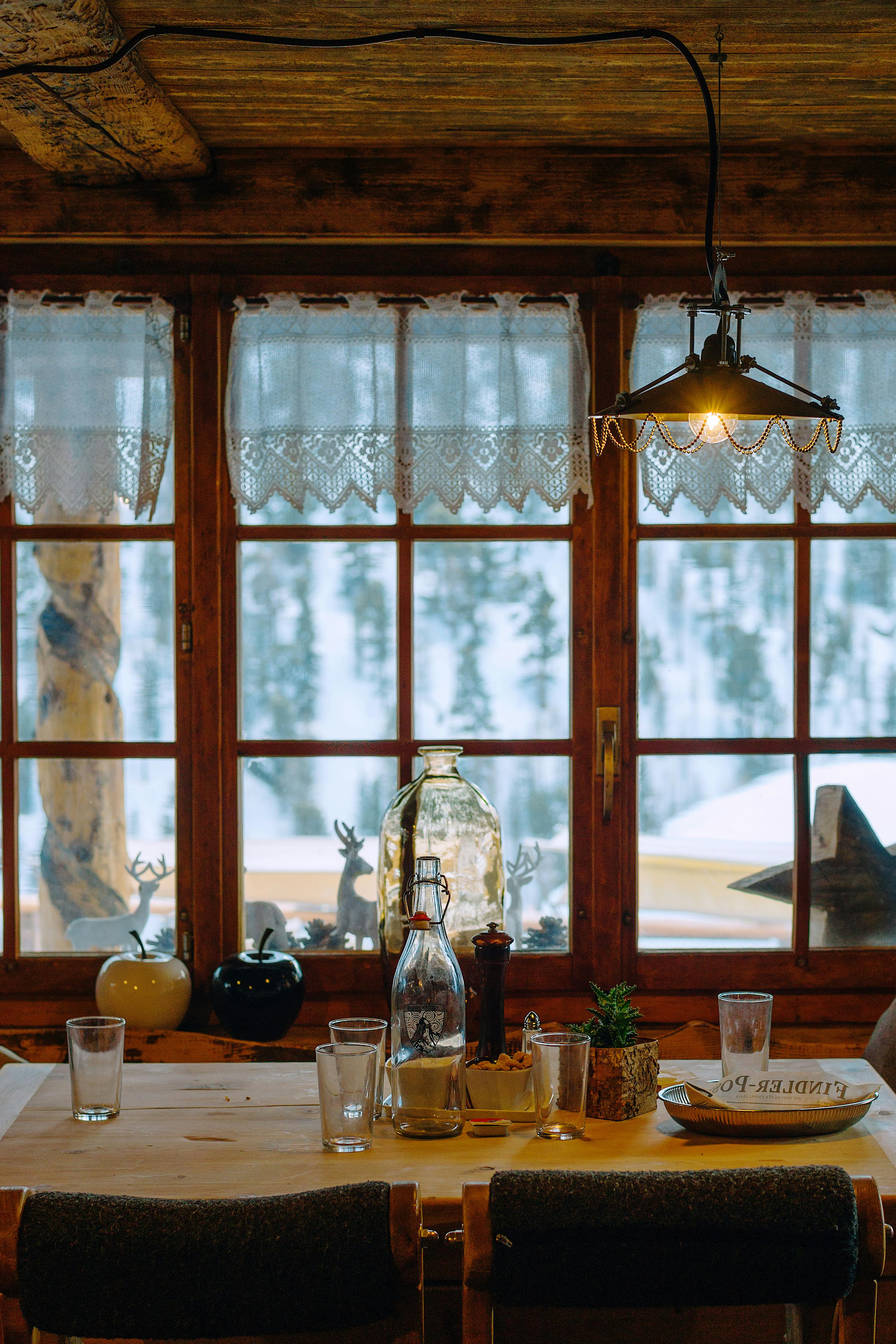 Cosy dining room with timber table, cabin-style decor, and large windows overlooking a snowy forest scene.
