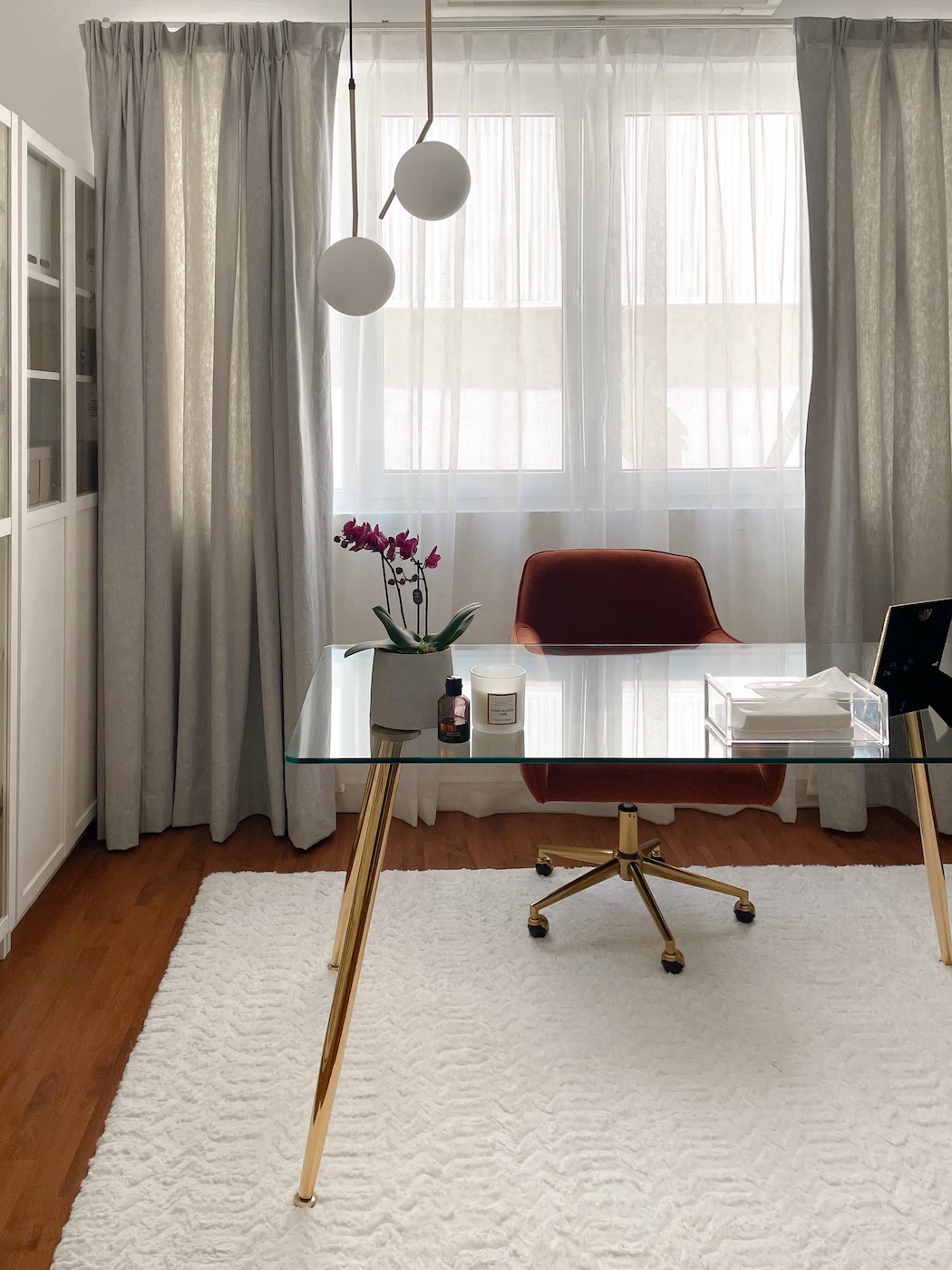 Elegant home office with glass desk, gold legs, velvet chair, pendant lights, and floor-to-ceiling curtains by Roberts Renovations.