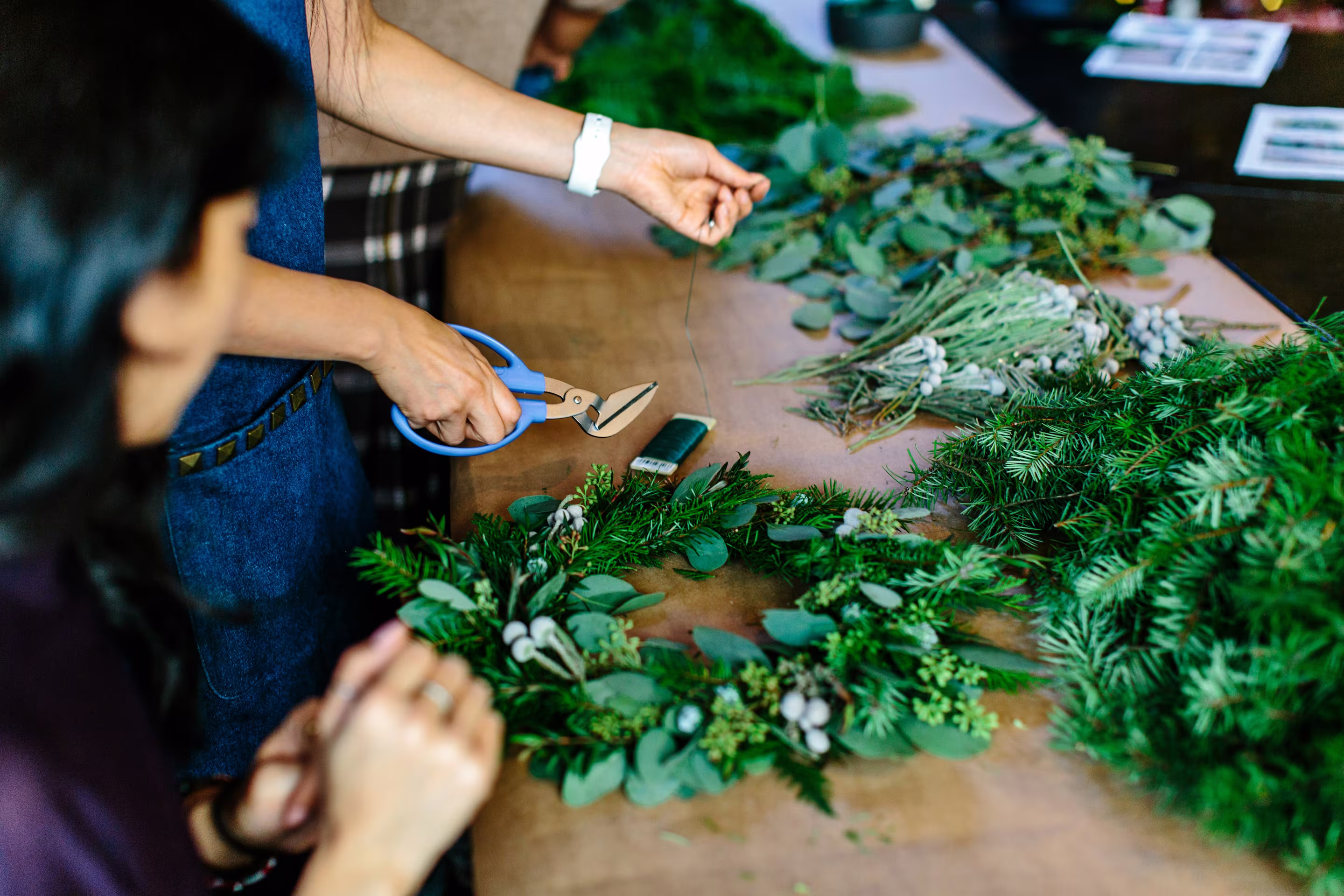 People creating a handmade Christmas wreath using fresh greenery, eucalyptus, and craft tools during a festive DIY workshop.