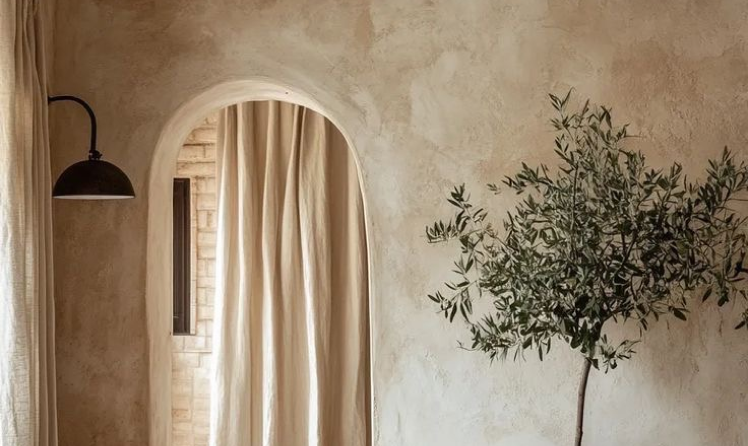 Soft, textured plaster wall with an arched doorway, flowing linen curtain, black metal sconce, and an olive tree adding organic warmth in natural light.