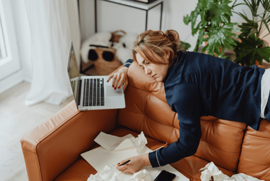 A lady lying n the back of her cuch surrounded by work and her laptop showing a state of burnout and work stress