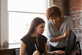 Two women are looking at a computer screen in an office setting. One woman is seated and looking at the screen while holding a pen. The other woman is standing behind her, leaning in and pointing at the screen.