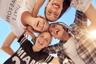 Four friends laugh together in a circle, looking down at the camera, against a bright blue sky.