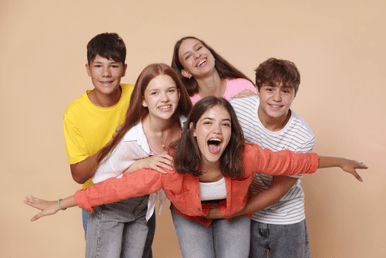 Five young people are posing closely together and smiling broadly against a peach-colored background. One person in the front is leaning forward with arms outstretched, while the others are behind, some with arms around each other.