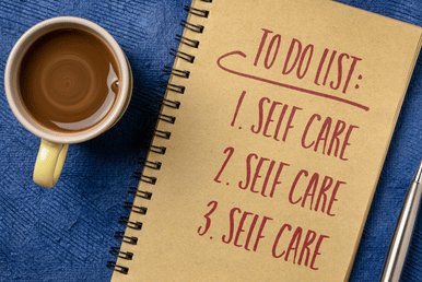 A notebook with "TO DO LIST" written on it, followed by "Self Care" listed three times, next to a cup of coffee and a pen.