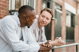 Two people talking while leaning on a railing. The person on the left is wearing a light blue shirt, and the person on the right has reddish-brown hair and is wearing a light beige shirt.