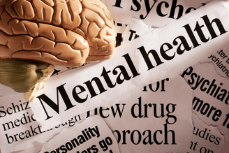 A model of a brain rests on torn newspaper clippings about mental health, including headlines about a new drug approach and psychiatric studies.