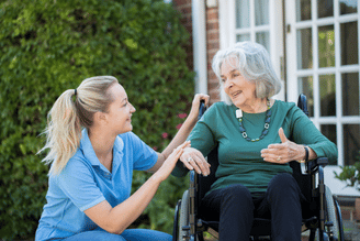 A caregiver in a blue shirt kneels next to an elderly woman in a wheelchair, both smiling and engaged in conversation outdoors.