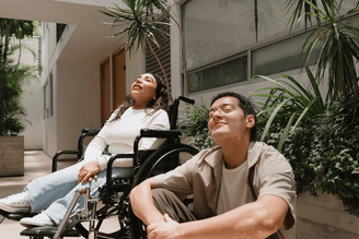Two people sitting outdoors, one in a wheelchair, both with their eyes closed and faces tilted towards the sun.