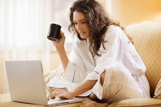 A person in a robe sits on a couch with a laptop and a mug.
