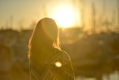 Back view of a person wearing a patterned sweater looking out at a bright sunset over a harbor.