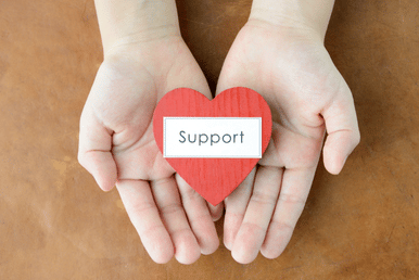 Hands holding a red heart with the word "Support" written on a white label.