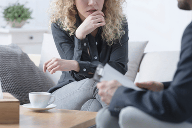 A person with curly blonde hair sits on a couch, looking concerned while resting their chin on their hand. In front of them, a person in a suit holds a clipboard. A coffee cup and tissue box are on a table between them.
