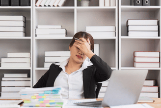 A person in a suit holds their forehead in frustration while sitting at a desk with a stack of papers and a laptop.