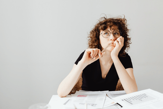 A person with curly hair and glasses sits at a desk with papers and a pen, looking thoughtful.