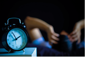 A close-up of an alarm clock on a nightstand with a person in the background tossing and turning in bed.