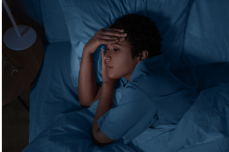 A person lies in bed at night with their hand on their forehead, appearing distressed or unable to sleep.