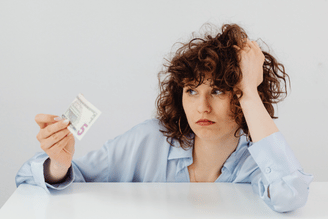 A person with curly hair holds a five-dollar bill in their hand, with their other hand resting on their head, looking away with a concerned expression.