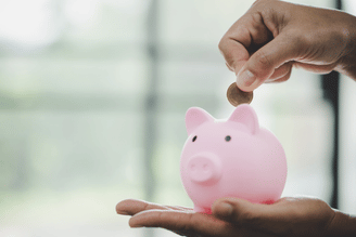 A hand holds a pink piggy bank while another hand inserts a coin into the slot.
