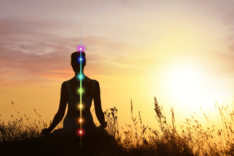 Silhouette of a person meditating outdoors at sunset with a visualization of the seven chakras glowing in rainbow colors along their spine.