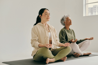 Two individuals are meditating in a yoga studio, sitting cross-legged on mats with their eyes closed and hands resting on their knees.
