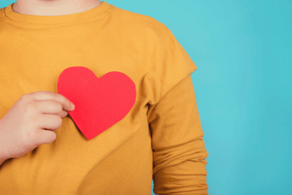A hand holds a red paper heart against a yellow sweater on a blue background.