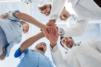 A group of medical professionals in scrubs and lab coats are seen from below, with their hands clasped together in a circle, symbolizing teamwork and unity.