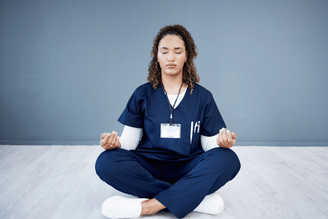A person in navy blue scrubs meditates cross-legged on a light wooden floor against a gray wall.