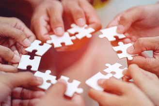 Several hands holding and connecting white puzzle pieces in a circular pattern.