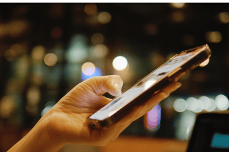 A hand holds a smartphone over a blurred nighttime city street with warm lights in the background, with the screen angled toward the viewer.