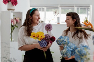 Two women in a bright flower shop smiling at each other while holding large bouquets of colorful flowers, with hanging flower displays and windows in the background.