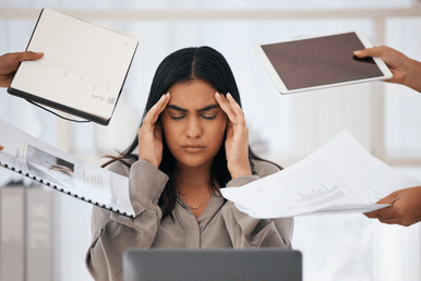 A stressed person sitting at a desk with hands on their head while multiple coworkers hold papers around them, suggesting being overwhelmed by work and deadlines.