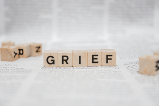 Scrabble tiles spelling “GRIEF” across a table of blurred documents, with additional letter tiles scattered at the sides.