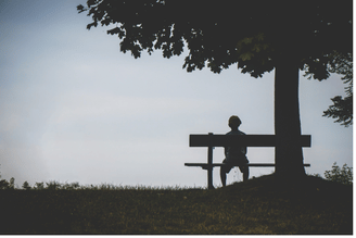 Silhouettes of two people sitting on a bench in a park near a large tree, with low grass and a hazy sky in the background.