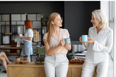 Two people in a bright office kitchen wearing white tops and holding blue mugs, standing and chatting near a counter with food and coffee supplies.
