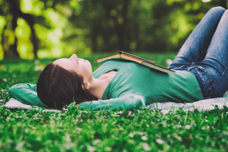 Two people lying on a grassy lawn in bright sunlight, one resting on the other’s chest while they hold hands, with trees and green foliage in the background.
