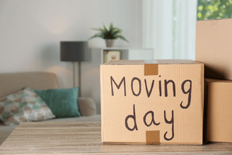 A cardboard sign on a wooden table reads “Moving day,” with stacked moving boxes nearby and a living room background with a sofa, cushions, and a small plant.